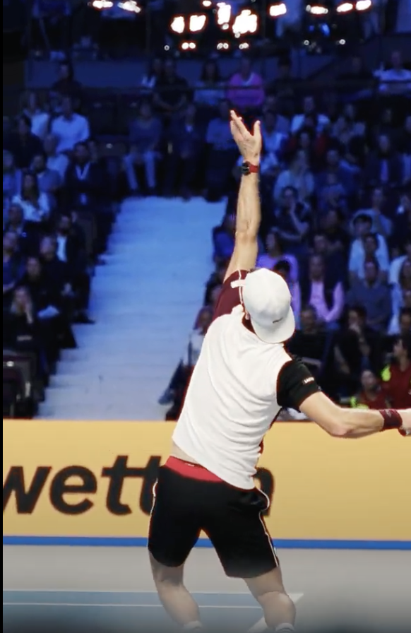 Ein Tennisspieler in sportlicher Kleidung, bestehend aus einem weißen Shirt, schwarzen Shorts und einer weißen Kappe, führt einen Aufschlag während eines Spiels aus. Der Spieler ist von hinten zu sehen, wie er sich mit ausgestrecktem Arm nach oben streckt, um den Ball zu schlagen. Im Hintergrund sind unscharf die Zuschauer auf den Tribünen zu erkennen, die das Spiel verfolgen. Die Szene ist dynamisch und fängt einen entscheidenden Moment im Tennismatch ein, während die Aufmerksamkeit des Publikums auf den Spieler gerichtet ist.