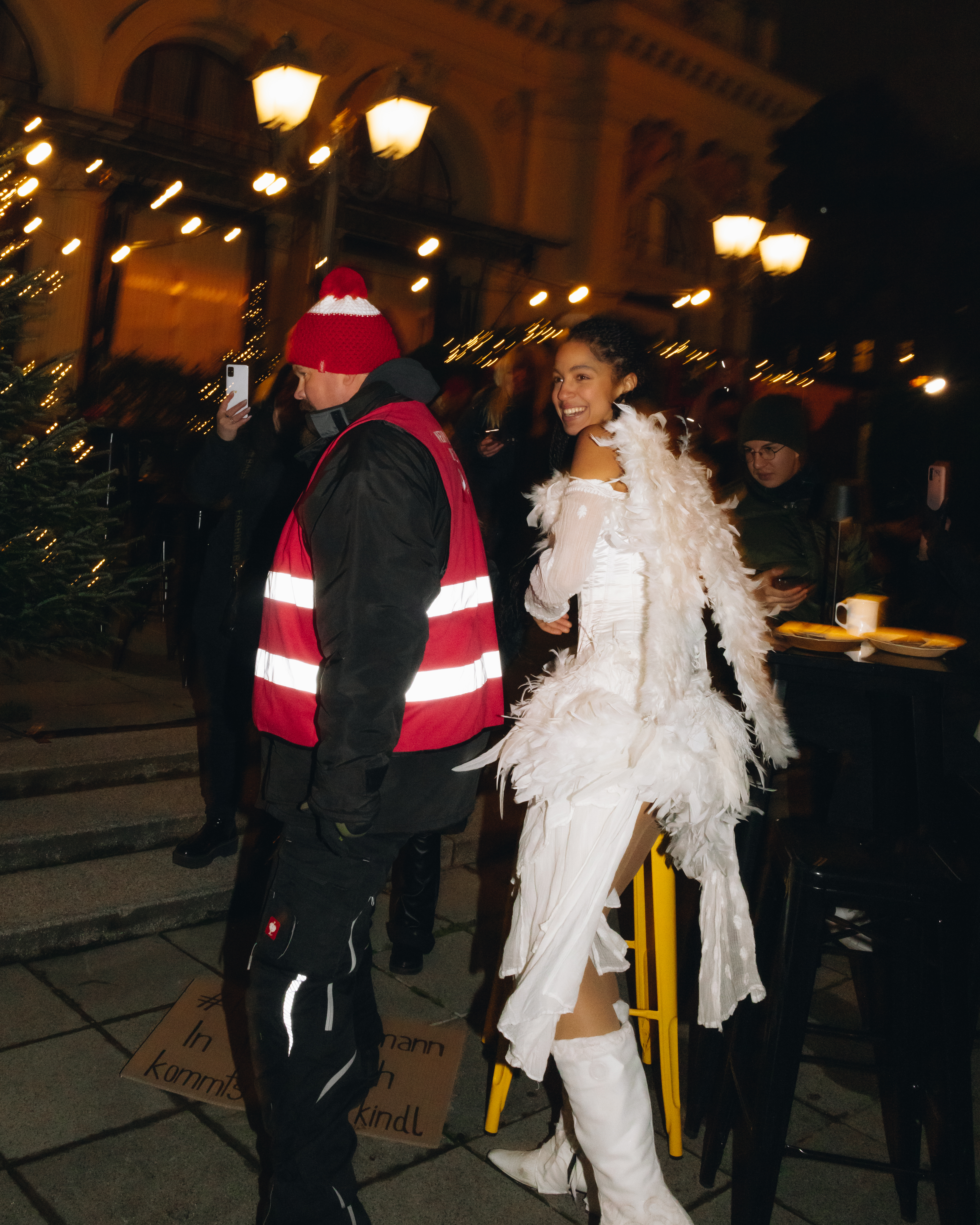 Eine junge Frau in einem weißen, engelhaften Kostüm mit Federn wird von Sicherheitskräften bei einer nächtlichen Veranstaltung im Freien abgeführt. Sie lächelt dennoch fröhlich in die Kamera, während ein Mann in einer roten Warnweste und Mütze neben ihr geht. Um sie herum sind Menschen in winterlicher Kleidung und im Hintergrund leuchten Lichter und weihnachtliche Dekorationen, die die Szenerie festlich erscheinen lassen. Trotz der Situation bleibt die Stimmung durch das Lächeln der Frau heiter und unbeschwert.