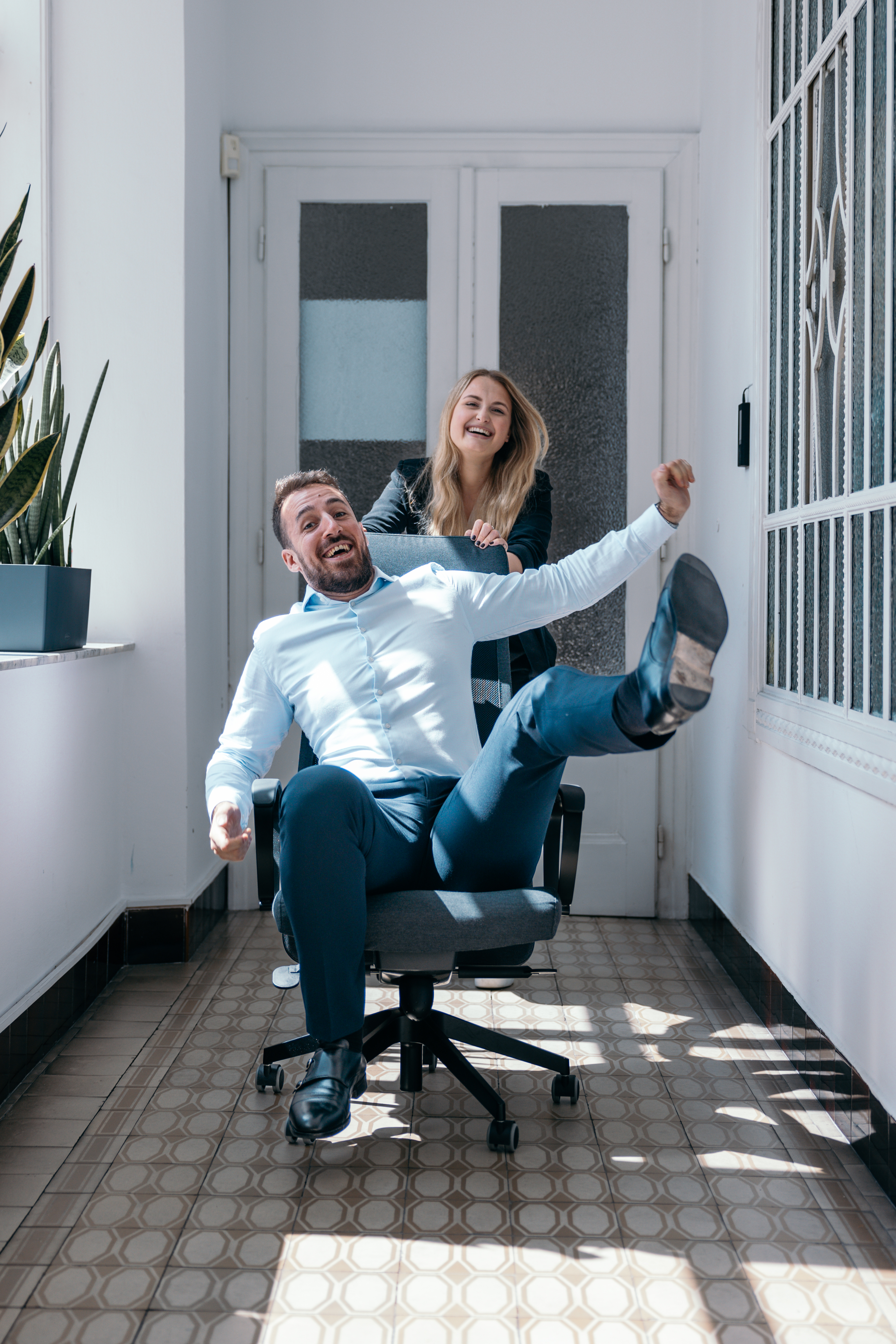 Ein Mann in einem weißen Hemd sitzt fröhlich in einem Bürostuhl und streckt ein Bein in die Luft, während eine Frau hinter ihm steht und den Stuhl schiebt. Beide lachen ausgelassen und scheinen viel Spaß zu haben. Sie befinden sich in einem hellen Flur mit Fliesenboden und Pflanzen am Fenster. Die Szene vermittelt eine spielerische und entspannte Atmosphäre im Arbeitsumfeld.
