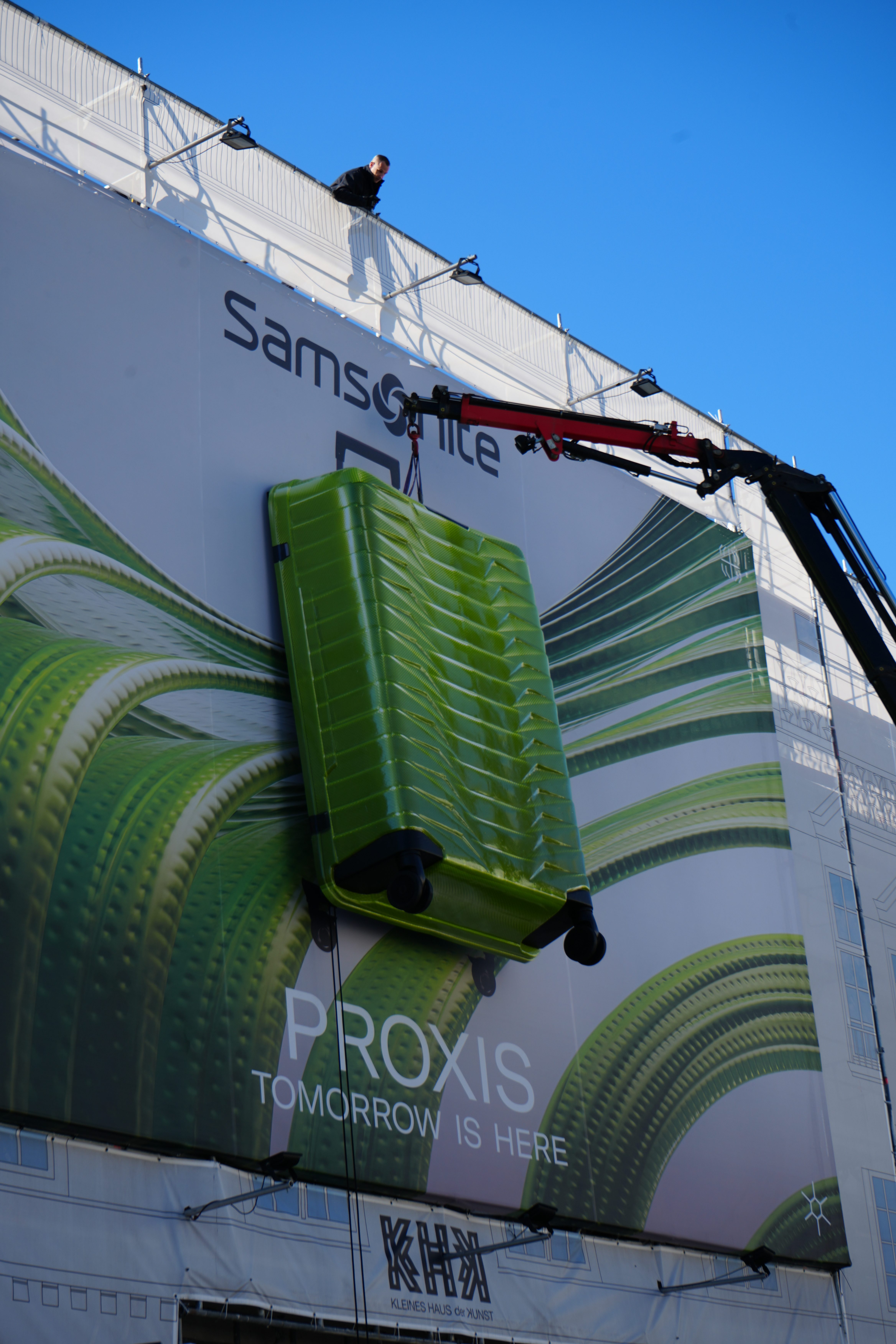 Ein großes Werbeplakat für die Marke Samsonite wird an der Fassade eines Gebäudes befestigt. Auf dem Plakat ist ein grüner Koffer zu sehen, der dreidimensional aus der Werbefläche herausragt. Ein Kran hebt den Koffer an, während ein Arbeiter oben auf dem Gebäude steht und das Geschehen überwacht. Die Werbung trägt den Text "PROXIS - TOMORROW IS HERE" und ist in einer modernen, futuristischen Ästhetik gestaltet. Der Himmel ist klar und blau, was die leuchtenden Farben des Plakats betont.